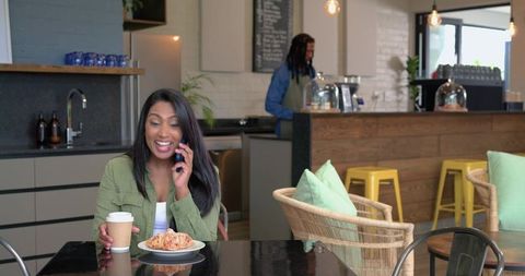 Smiling diverse woman talking on phone while holding coffee and croissant in modern cafe