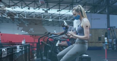 Woman wearing mask using phone on stationary bike in industrial gym workout