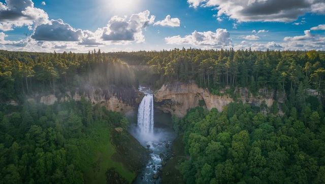 Sunlit waterfall plunging into misty canyon amid evergreen forest canopy (aerial)