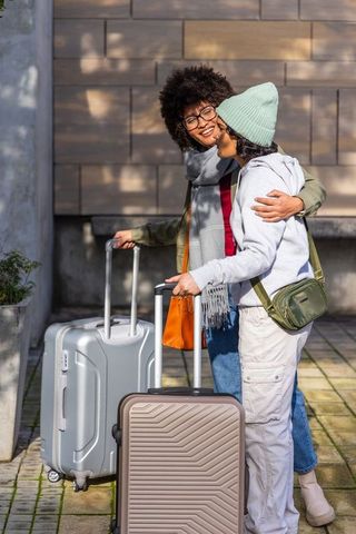 Diverse female friends hugging, smiling and holding rolling suitcases outside building