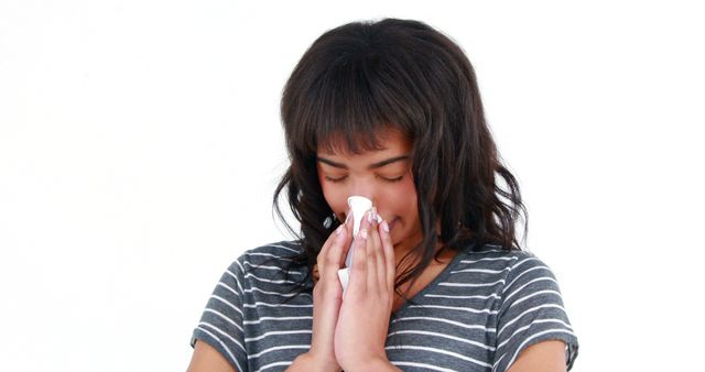 Young woman blowing her nose with tissue paper, indicating cold, flu, or seasonal allergies. She is wearing striped shirt, standing isolated against white background. Useful for articles on healthcare, wellness, hygiene practices, and medical advisories regarding colds and allergies.
