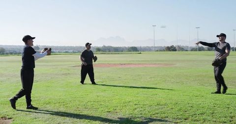 Three Male Teammates Practicing Baseball Throws on Sunny Grass Field with Dirt Infield and Caps