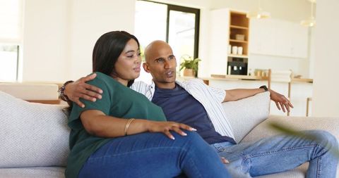 Diverse couple cuddling on sofa in bright modern open-plan living room, relaxed together