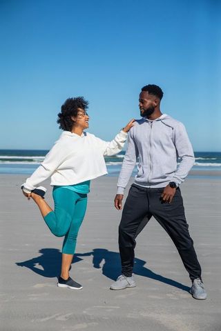 Diverse couple stretching quads on sandy beach in activewear