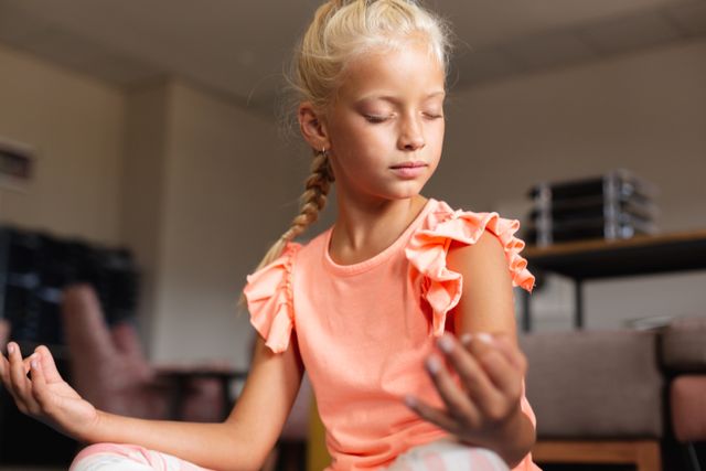 Young Caucasian schoolgirl practicing meditation and yoga in a classroom setting. Ideal for use in educational materials, wellness programs, children's activity guides, and mindfulness training resources. Highlights themes of childhood development, mental health, and physical fitness.