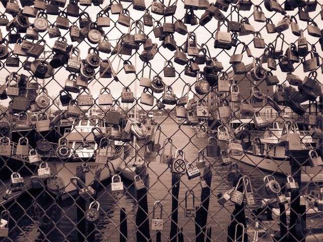 Love locks attached to a chain link fence near a waterfront area with boats visible in the background. Widely used as a symbol of unbreakable love and commitment by couples, this scene is ideal for use in themes related to romance, urban tradition, travel, and nautical settings. It conveys themes of love, partnership, and urban culture.