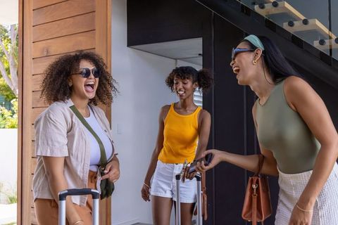 Diverse women laughing and arriving at modern home with rolling luggage and handbags