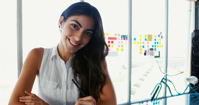 A young Caucasian woman smiles at the camera, holding a pen in a bright office environment, with copy space. Her cheerful demeanor suggests a positive work atmosphere, in a creative or startup setting.