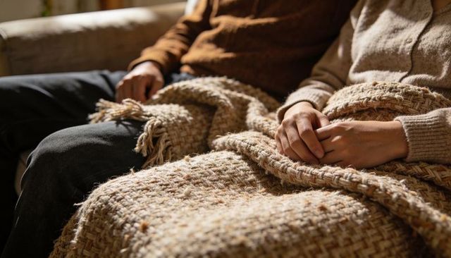 Couple cuddling under chunky knit blanket on sofa wearing cozy sweaters and warm tones
