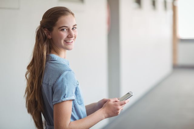 Young schoolgirl smiling while using a mobile phone in a school corridor. Ideal for educational content, technology in education, student lifestyle, and communication themes.