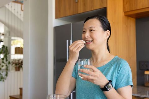 Mature asian woman taking supplement smiling holding glass of water in modern kitchen