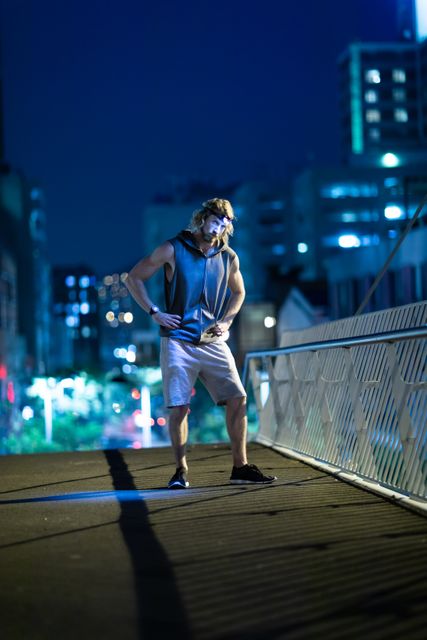 Front view of a fit Caucasian man with long blonde hair wearing sportswear exercising outdoors in the city at the evening, standing with hands on hips on the footbridge, wearing head light.