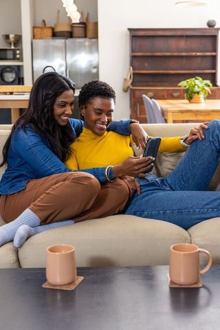 Indian and african american couple cuddling on beige sofa, smiling while viewing smartphone