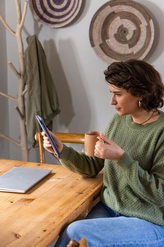 30s woman working from home holding tablet and mug at wooden table in cozy green sweater