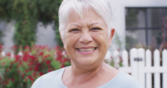 Senior woman with short gray hair smiling happily in an outdoor setting with a white picket fence and vibrant garden in the background. Ideal for depicting joy and positive aging, promotional materials for senior living, wellness content, or family-centric advertisements.