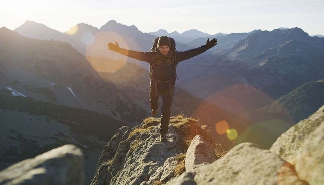 Hiker balancing on narrow alpine ridge at sunrise with backpack and lens flare