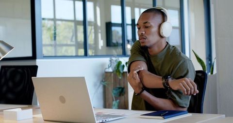 Young professional stretching while working at laptop with headphones in modern office