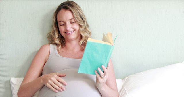 A young Caucasian pregnant woman is smiling while reading a book, comfortably seated against pillows, with copy space. Her relaxed demeanor and the book suggest she's enjoying a peaceful moment of leisure during her pregnancy.