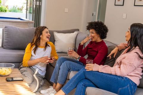 Laughing diverse friends lounging on living room sofa sharing drinks and snacks in cozy gathering