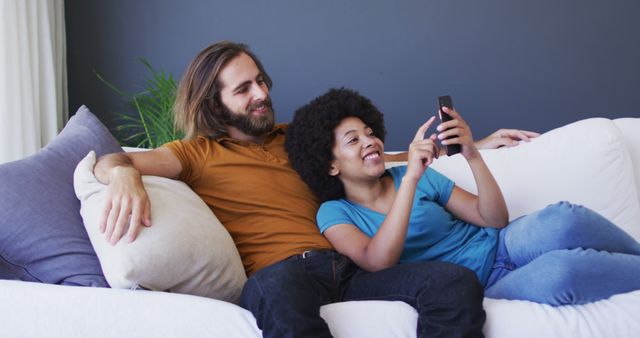 A multiracial couple relaxing together on a couch while using a smartphone. Ideal for blogs, articles, and advertisements focusing on relationships, happiness, technology, home life, and leisure activities. This image conveys feelings of comfort, connection, and modern living.
