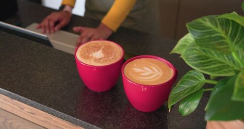 Female barista in mustard yellow leaning on stone counter with two red cups of latte art