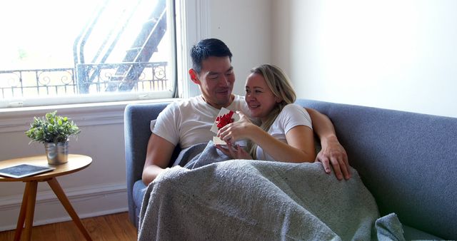 Depicts a happy couple relaxing on a sofa by a window, sharing a gift and smiling. They are wrapped in a blanket, creating a cozy atmosphere. Perfect for illustrating themes of love, togetherness, gifts, and indoor relaxation in home settings. Ideal for use in promotional materials for lifestyle, home decor, or relationship-related content.