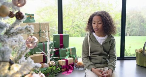 African American boy sitting on windowsill near Christmas tree and wrapped gifts