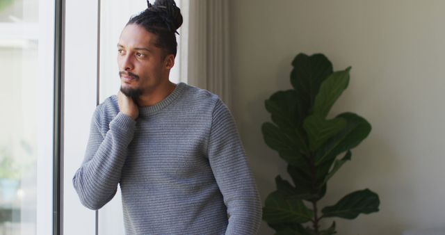 Man standing by window, wearing a gray sweater, leaning on his hand, looking thoughtful. Green plant adds to the cozy home ambiance. Useful in lifestyle blogs, mental health awareness content, reflective articles, or advertisements related to home goods and apparel.