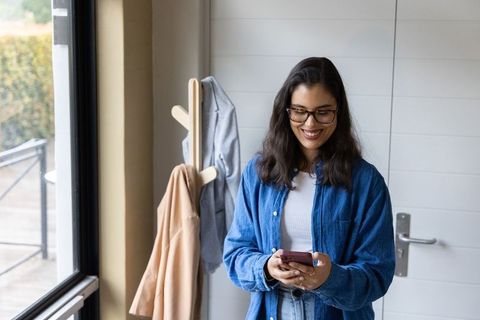 Hispanic woman smiling while checking smartphone by entryway window wearing denim shirt
