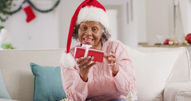 Senior woman wearing santa hat holding gift and smiling in living room. Ideal for themes related to Christmas celebrations, holiday excitement, elderly joy, and traditional holiday activities. Perfect for greeting cards, holiday advertisements, and festive marketing campaigns.