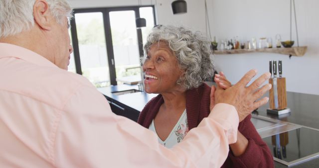 This joyful image shows a senior couple dancing and smiling in a modern kitchen. Ideal for use in content related to retirement lifestyle, senior romance, healthy aging, or family bonding. Perfect for websites, blogs, and advertisements promoting senior care, retirement communities, or family-centric products.