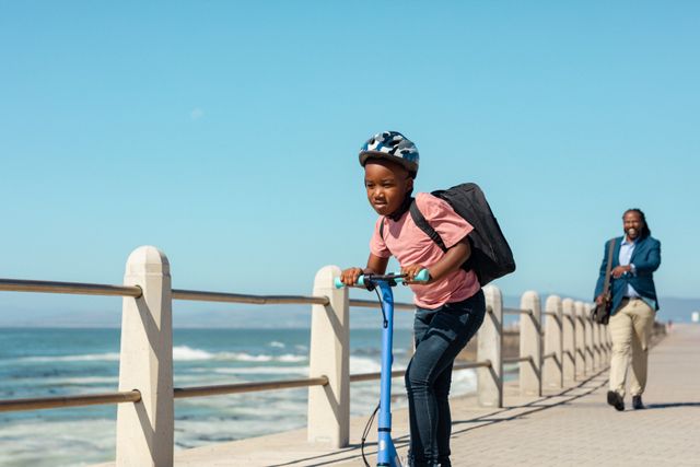 African american boy riding push scooter with father walking on promenade at beach during sunny day. unaltered, parenting, family, lifestyle and togetherness concept.