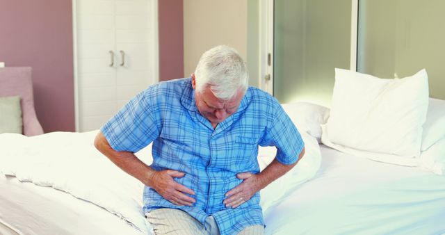 Elderly man sitting on bed holding stomach in pain, suggesting gut issues or other medical problems. Ideal for use in medical articles, healthcare advertisements, or senior care promotions.