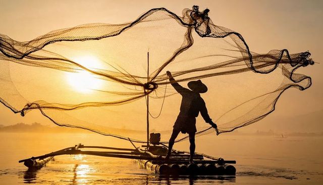 Throwing cast net from bamboo raft at sunrise silhouetting fisherman with conical hat