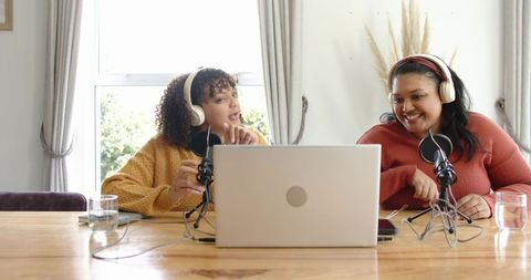 Female co-hosts recording home podcast at wooden table with laptop and microphones
