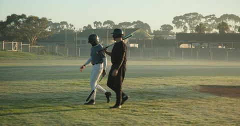 Morning baseball teammates walking across misty field carrying bats and helmets