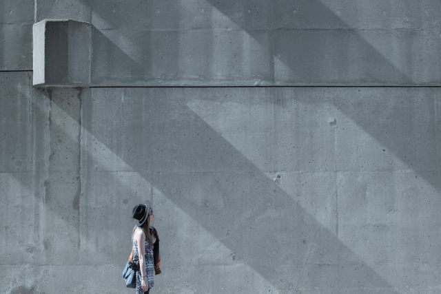 Woman looking up at a large concrete wall with dark shadows cast by beams. She wears casual street style clothing with a hat, suggesting a moment of contemplation or curiosity. This image can be used for urban lifestyle themes, introspection, solitary moments, or creative urban photography concepts.