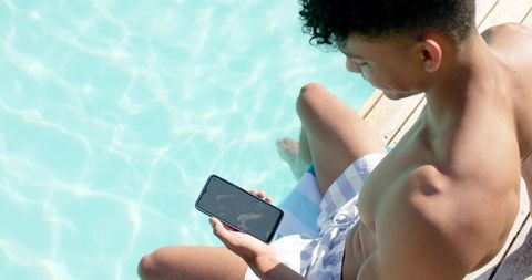 African american man in his twenties relaxing on pool deck using smartphone