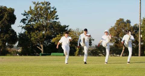 Cricket players performing synchronized high-kick warmups on grassy pitch in warm sunlight