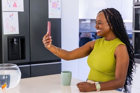 Smiling african american woman taking selfie in modern kitchen with fishbowl and smartwatch