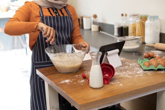 Woman in hijab mixing dough in a modern kitchen while using a tablet. Ideal for content related to cooking, baking, modern kitchen setups, inclusivity, and domestic life. Can be used in blogs, cooking websites, advertisements for kitchen gadgets, and lifestyle articles.