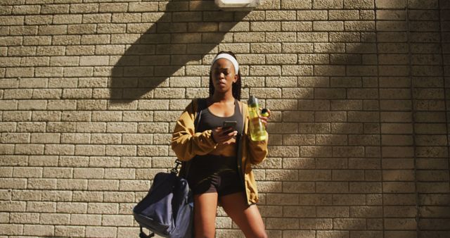 Young African American woman wearing athletic clothing standing against brick wall holding a phone in one hand and a water bottle in the other. She has a gym bag slung over her shoulder and wears sunglasses with a white headband. Suitable for promoting fitness, urban lifestyle, health, and exercise related content.