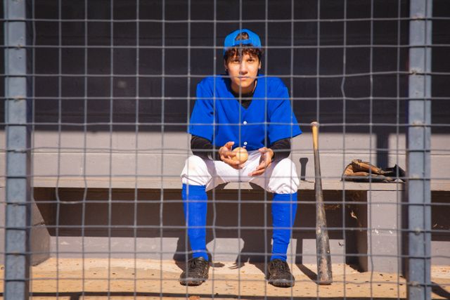 Female baseball player in blue uniform sitting on bench in dugout, holding baseball, with bat and glove nearby. Ideal for sports training materials, team spirit promotions, and athletic event advertisements.
