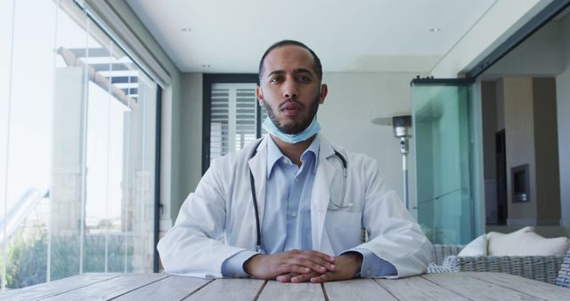 Young male doctor wearing white coat and stethoscope is sitting at a wooden table in a modern office environment. This image can be used for articles on healthcare, medical consultations, or professional medical settings. Ideal for hospital promotional materials, medical blogs, or healthcare employment resources.