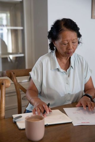 Senior asian woman reviewing finances at home office table with notebook and coffee mug