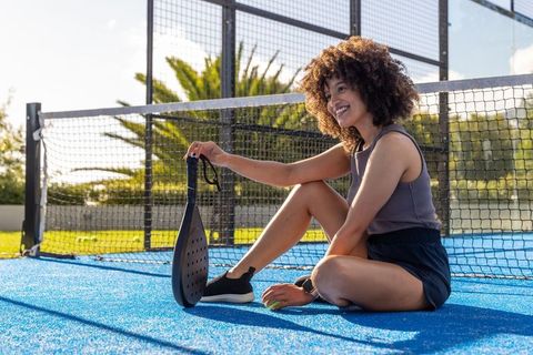Smiling african american woman sitting on blue padel court holding racket and ball