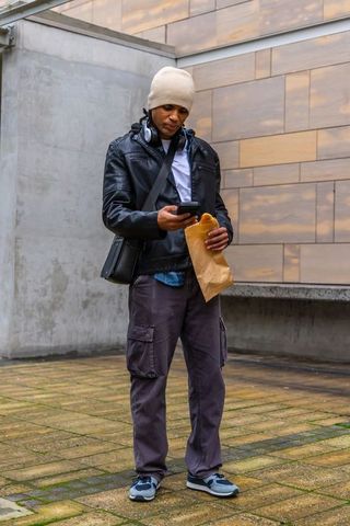 Standing man checking phone holding takeout in wet urban courtyard wearing leather jacket