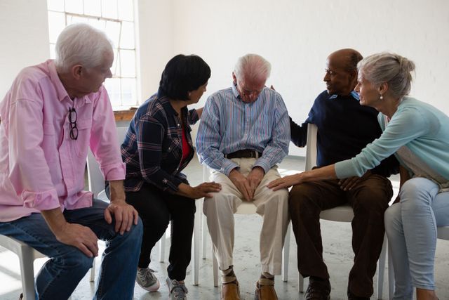 Senior friends are gathered around a male friend, offering comfort and support during a group discussion. This image can be used to depict themes of friendship, emotional support, community care, and mental health among elderly individuals. It is suitable for use in articles, advertisements, and campaigns related to senior care, mental health awareness, and community support programs.