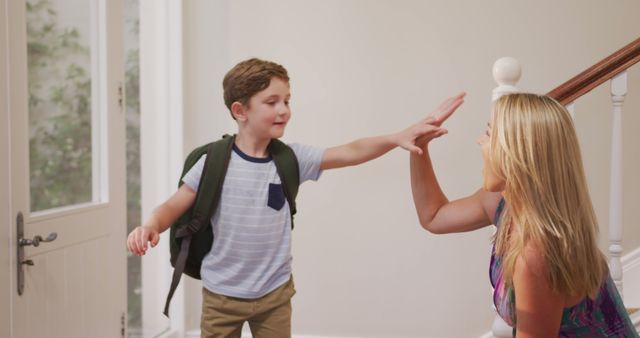 Young boy giving high-five to his mother in entrance hallway before leaving for school. Both smiling and appearing cheerful, creating a warm and loving family scene. Can be used for themes around family moments, school mornings, parenting, education, bonding, and enthusiasm for school.