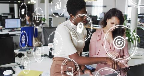Female Colleagues Collaborating on Tablet with Digital Interface Icons in Open-Plan Office